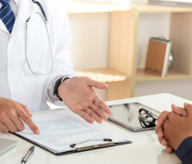 Doctor discussing health insurance or medical coverage with a patient during an office consultation, with paperwork and a clipboard on the desk.