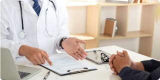 Doctor discussing health insurance or medical coverage with a patient during an office consultation, with paperwork and a clipboard on the desk.