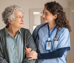 A healthcare worker in blue scrubs gently supports an older woman as they smile at each other in a bright home setting.