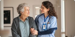 A healthcare worker in blue scrubs gently supports an older woman as they smile at each other in a bright home setting.