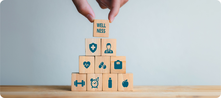 A hand places a wooden block labeled “WELLNESS” atop a pyramid of blocks featuring healthcare and healthy lifestyle icons.